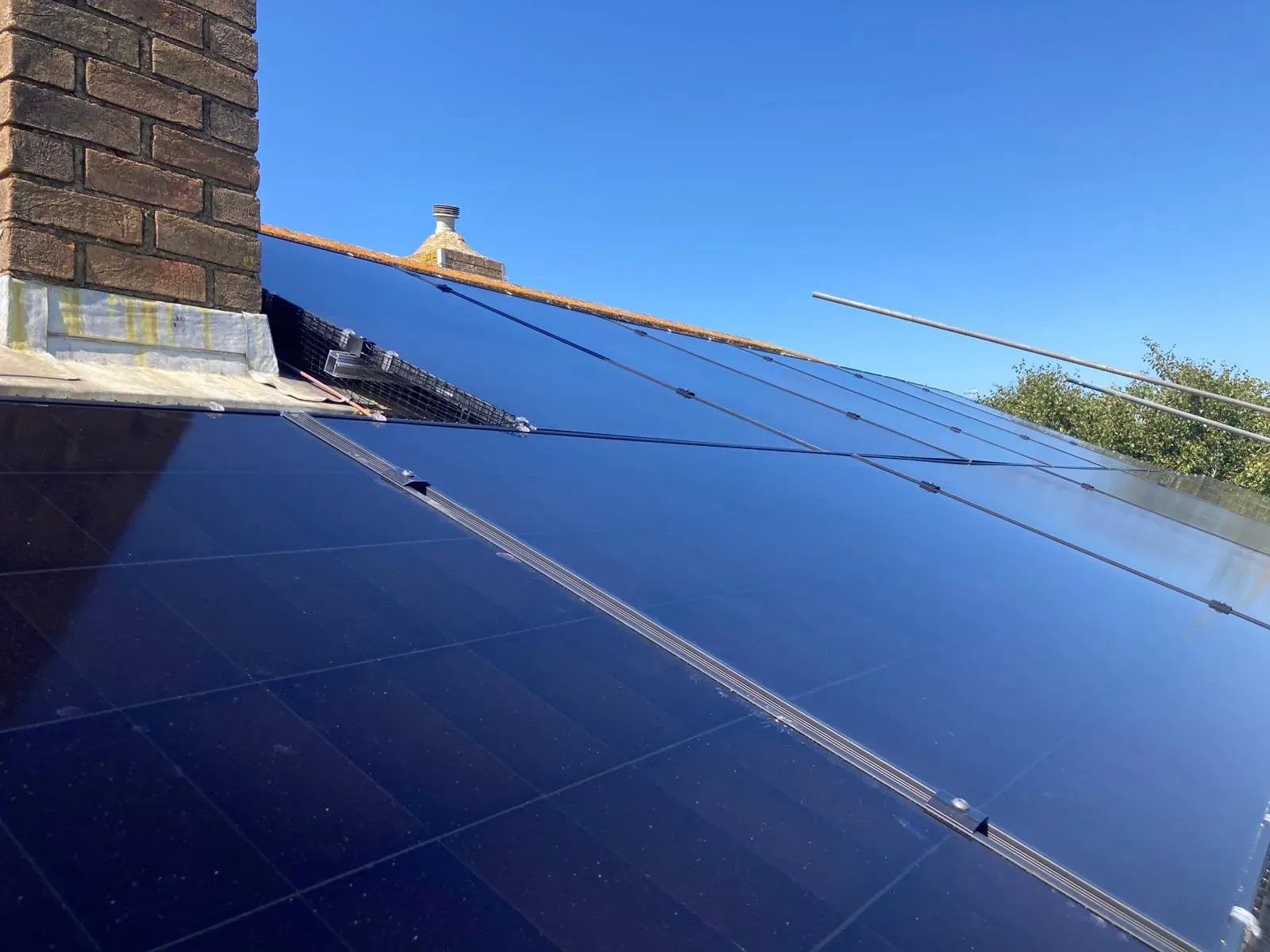 A close-up of solar panels on a tiled roof with a chimney behind, showing the mounting rails and clamps